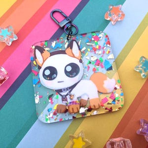 May include: A square keychain featuring a cartoon fox with large eyes, brown and white fur, and a small collar. The keychain is clear with colorful confetti and a gunmetal clasp. Decorative star-shaped embellishments surround the keychain.