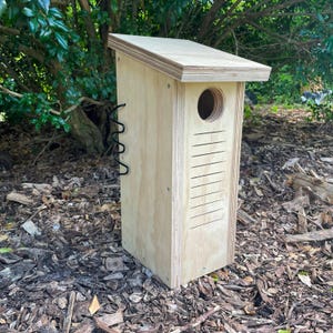 May include: A wooden birdhouse with a sloping roof, a circular entrance hole, and ventilation slits. The birdhouse is made of light-coloured wood and has a black metal hanging loop. It is set outdoors on a bed of mulch and wood chips.
