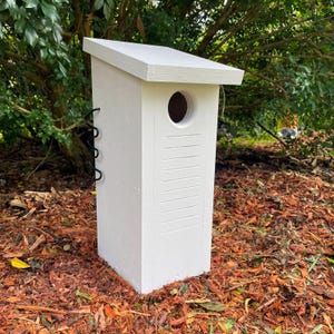 May include: A white wooden birdhouse with a slanted roof and a circular entrance hole. The birdhouse has vertical lines on the front and a black metal hanging loop. The birdhouse is set on a bed of brown mulch.