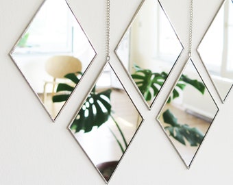 May include: Five diamond-shaped mirrors hanging on chains against a white wall. The mirrors reflect a room with a chair and a large-leafed plant. The mirrors have a silver frame.
