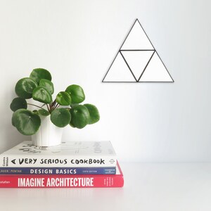 May include: A stack of three books on a white surface. The top book is white with black text that reads "A Very Serious Cookbook". The middle book is blue with white text that reads "Design Basics Sixth Edition". The bottom book is red with white text that reads "Imagine Architecture". A small green plant is in a white pot to the left of the books. A black geometric triangle wall decor is on the white wall behind the books.