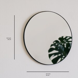 May include: A round black framed mirror with a diameter of 9.5 inches (24 cm). The mirror is reflecting a green plant.