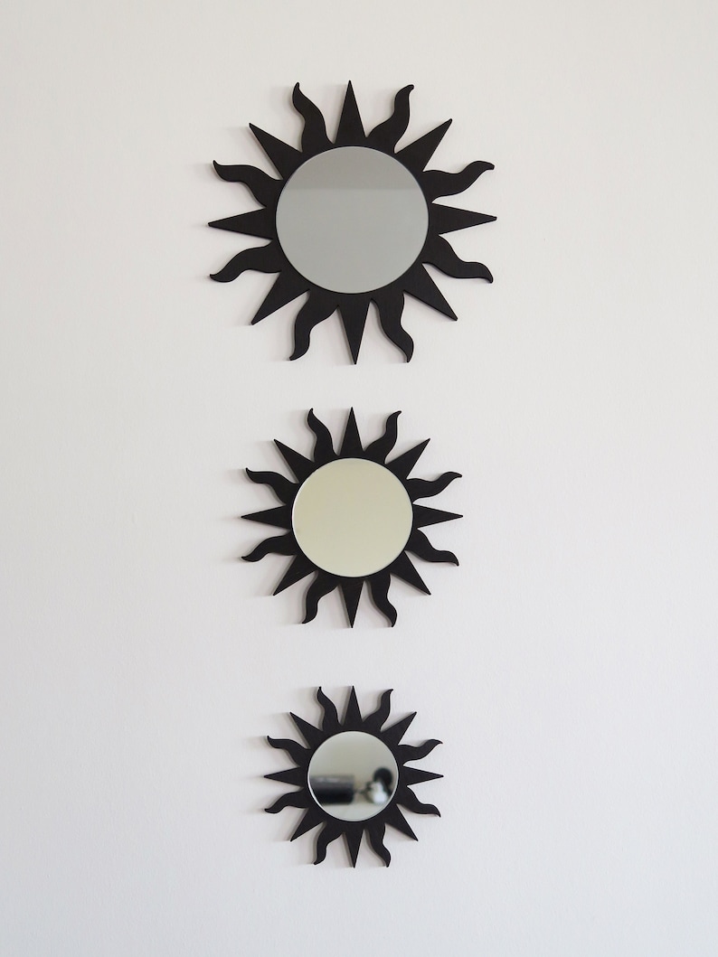 May include: Three black sunburst mirrors of different sizes are hanging on a white wall. The mirrors are round and have a black frame with a sunburst design. The mirrors are arranged vertically, with the largest mirror at the top and the smallest mirror at the bottom.