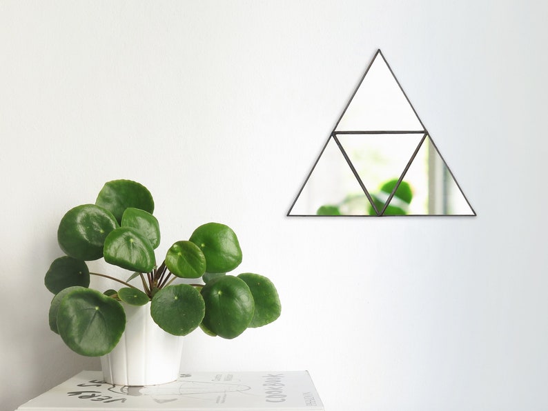May include: A triangular mirror with a black frame, divided into four smaller triangles. The mirror is hanging on a white wall, with a green plant in a white pot in the foreground.