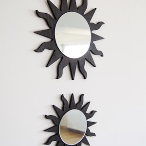 May include: Two black sunburst mirrors with round reflective surfaces. The larger mirror is above the smaller mirror.