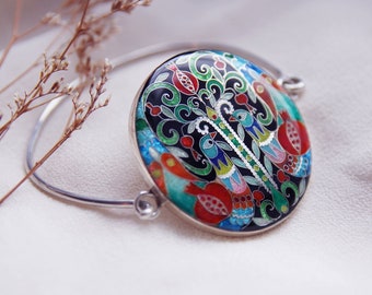 Puede incluir: Un brazalete de plata con un dije redondo de esmalte que presenta un diseño colorido de árbol de la vida con pájaros y granadas.