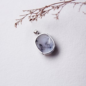 May include: A silver pendant with a white and gray oval-shaped stone. The stone has a cloudy, opaque appearance with black flecks throughout.