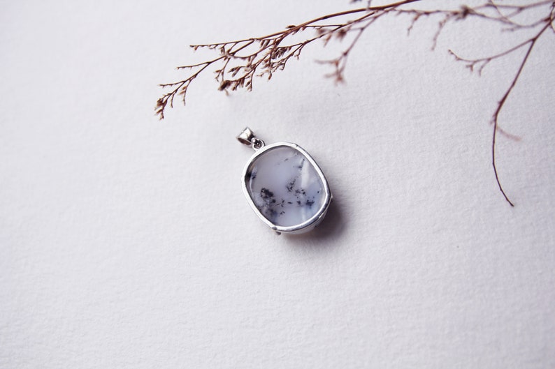 May include: A silver pendant with a white and gray oval-shaped stone. The stone has a cloudy, opaque appearance with black flecks throughout.