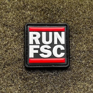 May include: A square, black patch with the white text "RUN FSC" and a red stripe. The patch is on a textured, olive green surface.
