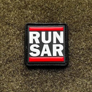 May include: A square patch with the words "RUN SAR" in white, bold letters. The text is set against a black background with red horizontal bars above and below the text. The patch is on a textured, olive-green surface.