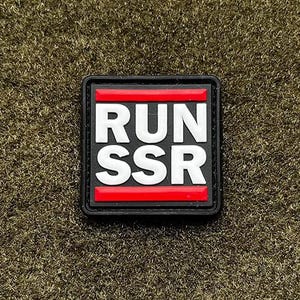 May include: A square patch with the text "RUN SSR" in white block letters on a black background. The letters are stacked vertically. A red stripe is above and below the text. The patch is on a textured, olive green surface.