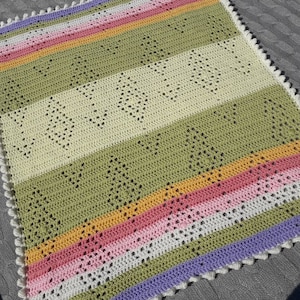 Puede incluir: Una manta de bebé de ganchillo con un patrón de rayas coloridas en tonos de verde, rosa, amarillo y morado. La manta tiene un borde festoneado y está hecha con un delicado patrón de punto abierto.