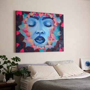 May include: A bedroom with a bed, two pillows, and a painting of a woman's face with her eyes closed. The painting is surrounded by colorful shapes and is hanging on a white wall. There is a plant on a side table next to the bed.