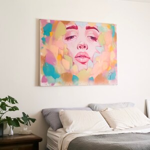 May include: A colorful abstract painting of a woman's face with a pink, blue, and yellow background. The painting is hanging above a gray bed with white pillows.