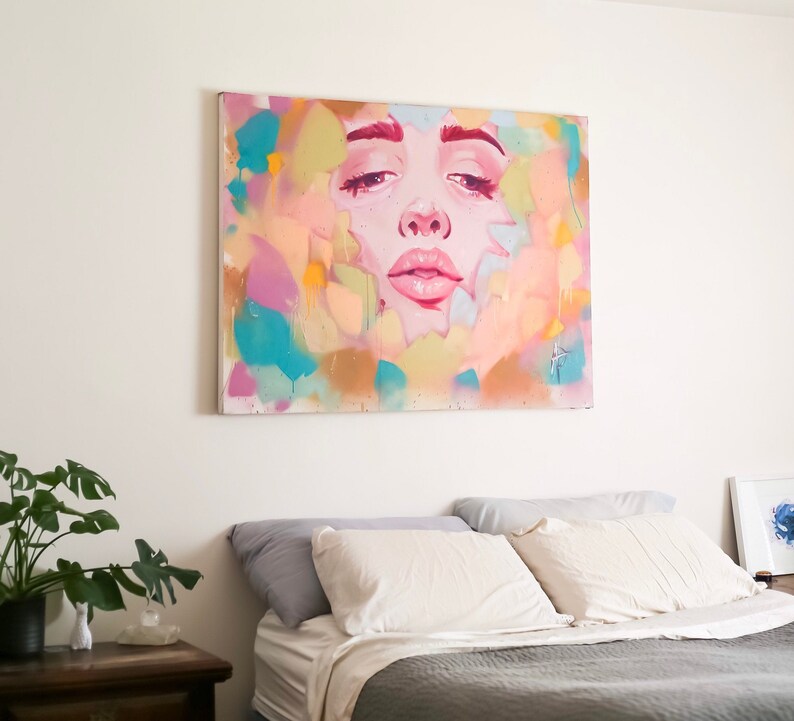 Puede incluir: Una pintura abstracta colorida de la cara de una mujer con un fondo rosa, azul y amarillo. La pintura est&aacute; colgada sobre una cama gris con almohadas blancas.