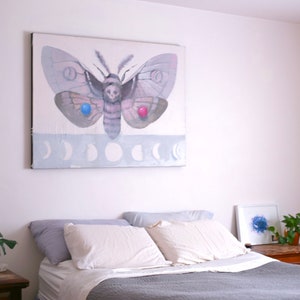 May include: A bedroom with a gray bedspread and white pillows. A painting of a moth with a skull on its head hangs on the wall above the bed. The moth has blue and red accents on its wings.