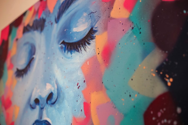 May include: Close-up of a colorful abstract painting featuring a woman's face with closed eyes. The painting is done in shades of blue, pink, yellow, and green, with splashes of white paint.