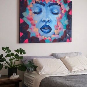 May include: A painting of a woman's face with closed eyes, surrounded by colorful abstract shapes. The painting is on a canvas and is hanging on a white wall above a bed with white pillows and a gray blanket.