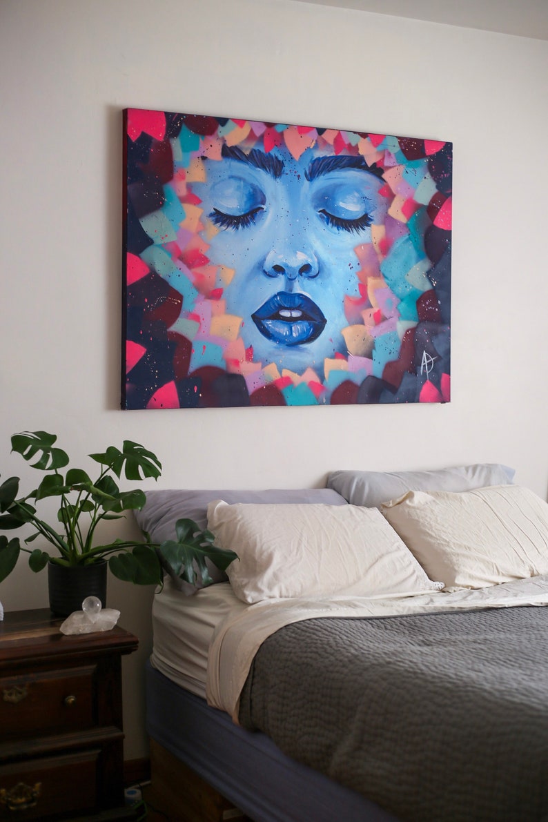 May include: A painting of a woman's face with closed eyes, surrounded by colorful abstract shapes. The painting is on a canvas and is hanging on a white wall above a bed with white pillows and a gray blanket.