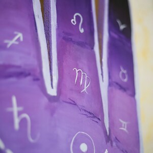 Puede incluir: Primer plano de una superficie pintada de p&uacute;rpura con s&iacute;mbolos del zod&iacute;aco blancos. El s&iacute;mbolo de Virgo es visible en el centro de la imagen.