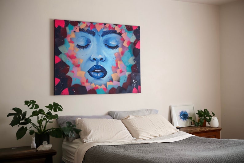 May include: A bedroom with a bed, two pillows, and a painting of a woman's face with her eyes closed. The painting is surrounded by colorful shapes and is hanging on a white wall. There is a plant on a side table next to the bed.