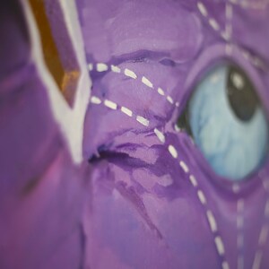 Puede incluir: Primer plano de un ojo pintado de morado con un contorno cosido blanco y un iris azul. El ojo es parte de una pintura m&aacute;s grande.