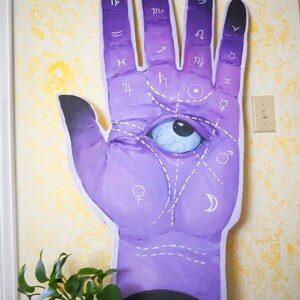 Puede incluir: Una mano morada con un ojo que todo lo ve pintado en la palma. La mano tiene s&iacute;mbolos astrol&oacute;gicos en cada dedo y l&iacute;neas dibujadas en la palma. El fondo es una pared amarilla.