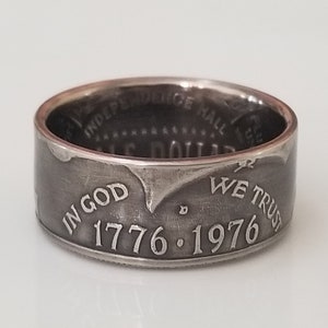 Può includere: Un anello d'argento con la scritta "IN GOD WE TRUST" e le date "1776-1976" incise sulla fascia. L'anello sembra essere fatto da una moneta, con le parole "INDEPENDENCE HALL" visibili all'interno.