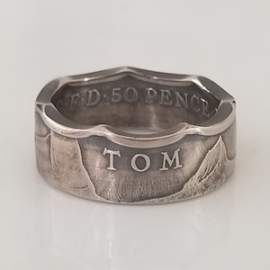 May include: A silver ring with the name "TOM" engraved on the front. The ring is made from a 50 pence coin, with the text "FD 50 PENCE" visible on the top edge. The ring has a textured, worn appearance.