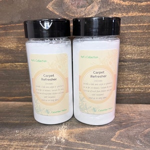 May include: Two white plastic bottles with black lids labeled "Kat's Collection Carpet Refresher" with the scent "Cucumber Melon". The bottles contain a white powder and are on a wooden surface.