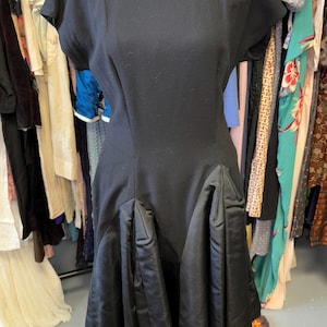 Op de afbeelding: Een zwarte vintage jurk met een getailleerd bovenstuk en een volle rok. De rok heeft een gerimpeld ontwerp en een gladde, satijnachtige textuur.