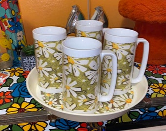 May include: A set of four white mugs with large handles, featuring a daisy pattern on a green background, resting on a matching tray. The mugs and tray have a retro aesthetic, with a white rim.