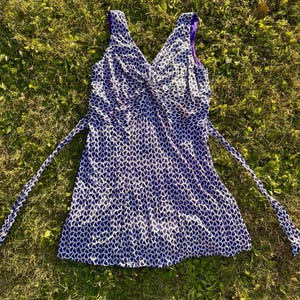 May include: A sleeveless dress featuring a deep V-neckline with a central twist detail. The garment is patterned with a repeating white and dark blue geometric heart-like print and includes two long side ties at the waist with a solid purple lining.