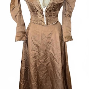 May include: A vintage brown dress with a high collar and long sleeves. The dress features a fitted bodice with decorative embroidery and a full skirt. The sleeves have a puffed design at the shoulders. The dress is made of a shiny fabric.