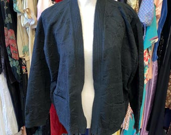 May include: A black open-front jacket with a textured pattern and two front pockets. The jacket is displayed on a mannequin, showcasing its design and fit. The garment is part of a clothing collection.