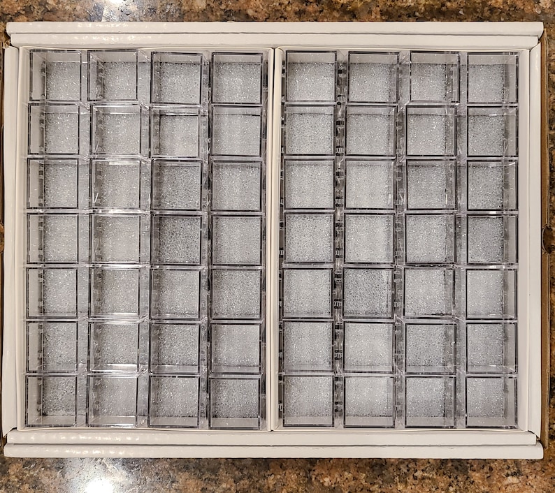 May include: A white cardboard box with two rows of clear plastic square compartments. Each compartment contains a gray, textured material. The box is designed for organizing small items, such as beads or jewelry.