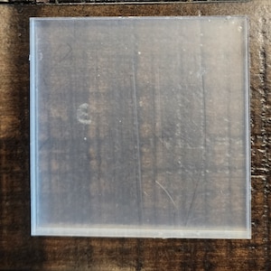 May include: A square, clear, semi-transparent acrylic sheet with smooth edges. The surface of the flat, rigid panel shows faint, thin, linear scratches and a small, light, circular mark near the left side, indicating a used condition.