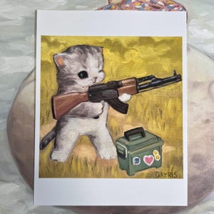 May include: An oil painting print on paper featuring a grey and white kitten standing in a yellow field, holding a black and brown rifle. Beside the kitten is a green ammunition box decorated with three small stickers, signed Dayris.