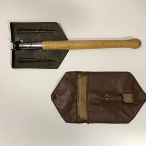 Puede incluir: Una pala plegable vintage con mango de madera y hoja de metal oscuro. La pala va acompañada de una funda de transporte de cuero marrón con cierre de botón a presión. Diseñada para cavar y otras tareas al aire libre.