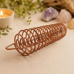 May include: A copper-colored metal coil sculpture featuring a cylindrical spiral design with a triangular loop at one end. The object is constructed from a single continuous wire formed into multiple concentric circles, creating a hollow tubular shape.