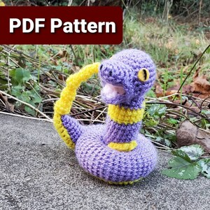 May include: Crocheted purple and yellow snake amigurumi toy. The snake has a yellow tail and a yellow stripe around its neck. The snake is sitting on a gray surface.