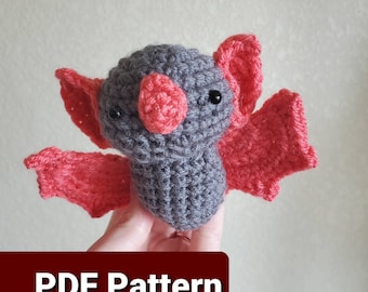 Puede incluir: Un juguete de amigurumi de murciélago tejido a crochet con cuerpo gris y alas rosas. El murciélago tiene ojos negros y una nariz rosa. El juguete está siendo sostenido por una mano. El texto "PDF Pattern" es visible en la parte inferior de la imagen.