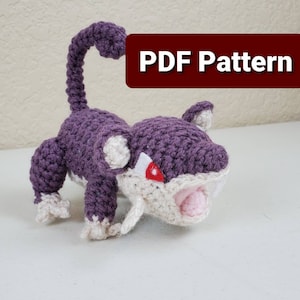 May include: Crocheted purple and white amigurumi toy of a Pokemon character with a red eye and a large mouth. The toy is posed in a standing position with its tail curled up. The text "PDF Pattern" is visible in the image.