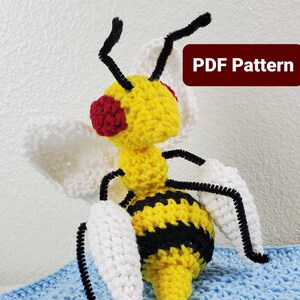 Puede incluir: Juguete de amigurumi de abeja de crochet con rayas amarillas y negras, alas blancas y ojos rojos. La abeja está sentada sobre una superficie azul. Patrón PDF.