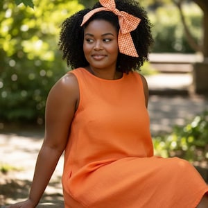 May include: An orange sleeveless dress with a matching gingham bow headband. The dress is knee-length and appears to be made of linen. The woman is sitting on a wooden bench outdoors, with a blurred green background.