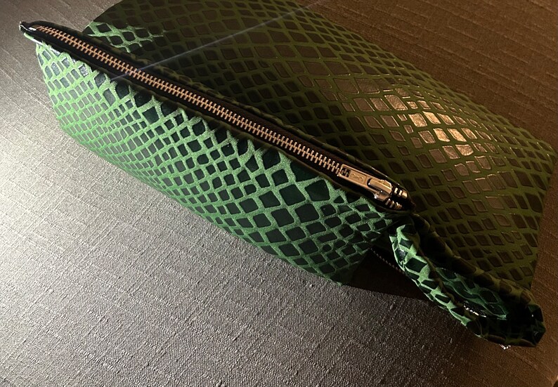 Pu&ograve; includere: Una pochette verde con cerniera a forma di pelle di serpente e una cerniera argentata.