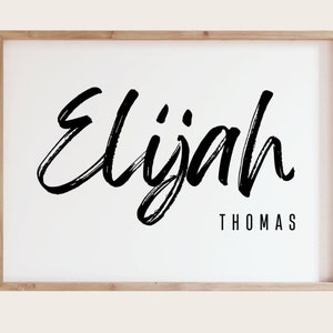 May include: A framed print featuring the name "Elijah" in bold, black, brushstroke lettering. Beneath, in a smaller, block font, is the name "Thomas". The frame is light wood, set against a white background.