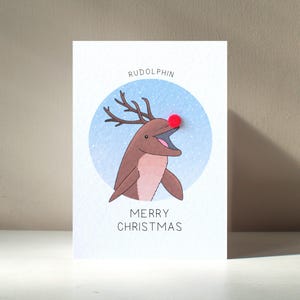 Biglietto di auguri natalizio personalizzato illustrato "Rudolphin" in formato A6 con personaggio Rudolph, delfino e renna, di Animixtures, con gioco di parole – Beneficia l'ente benefico