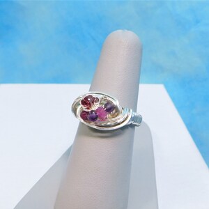 Puede incluir: Un anillo artesanal de alambre de plata con un grupo de pequeñas y vibrantes piedras preciosas. El anillo presenta un diseño de alambre tejido, con una disposición central de piedras rojas y moradas. El anillo se exhibe sobre un soporte gris.