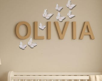 May include: A wall decoration featuring the name "OLIVIA" in large, tan-colored letters. Delicate white butterflies with silver accents are scattered around the name. The scene is set against a neutral-toned wall, with a white crib visible below.